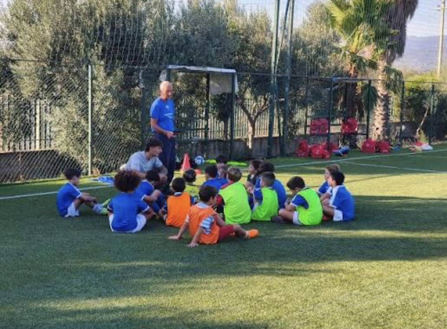 Angelo Orlando in a blue shirt leading a team talk with young players